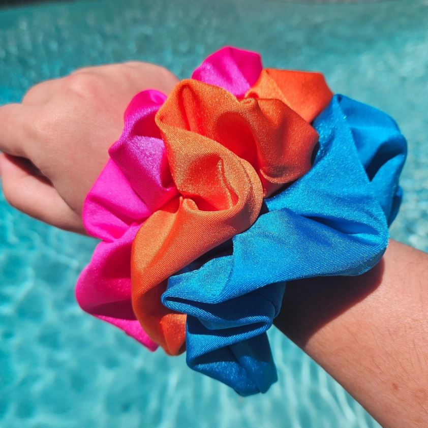 Bright pink, orange, and blue swim scrunchies worn on a wrist beside a swimming pool.
