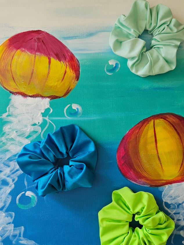 Mint green, teal blue, and neon green swim scrunchies arranged on a colorful jellyfish painting.