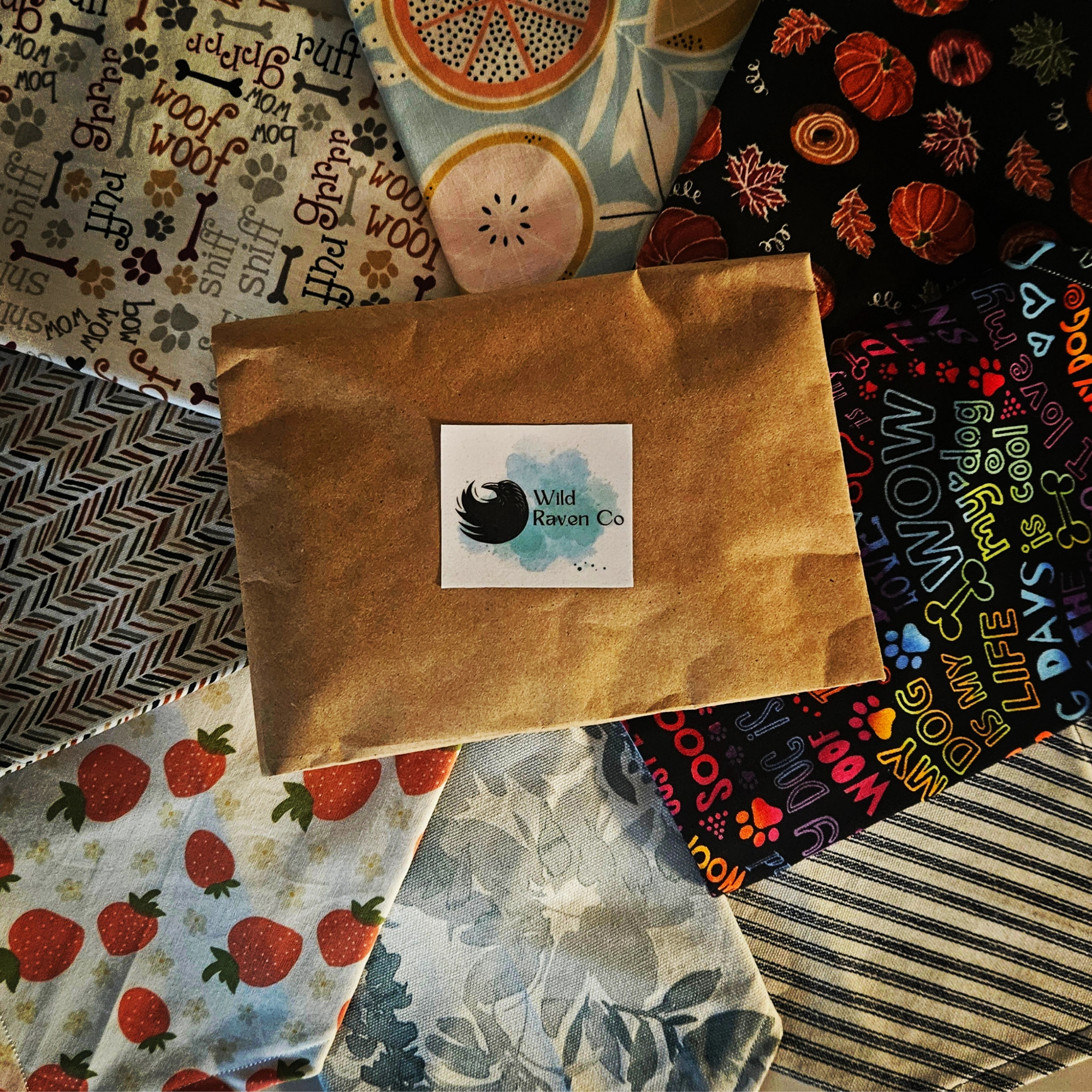 Wild Raven Co. mystery bandana bundle packaged in a kraft envelope and surrounded by assorted patterned fabric bandanas.
